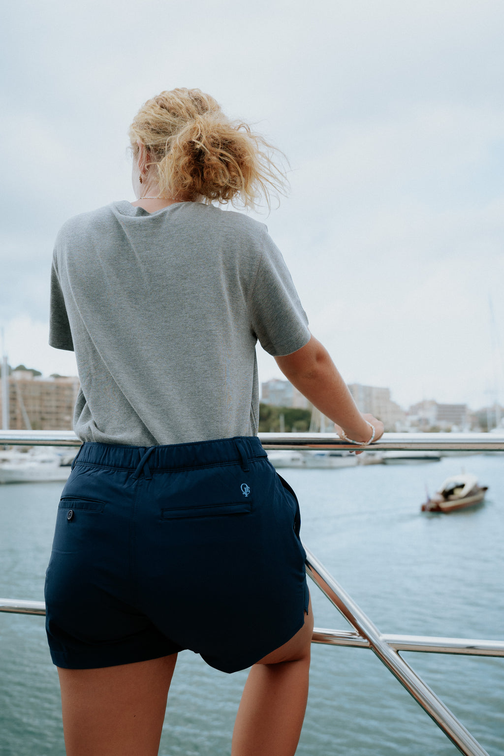 Woman wearing navy tech deck shorts standing on a boat overlooking water and buildings.
