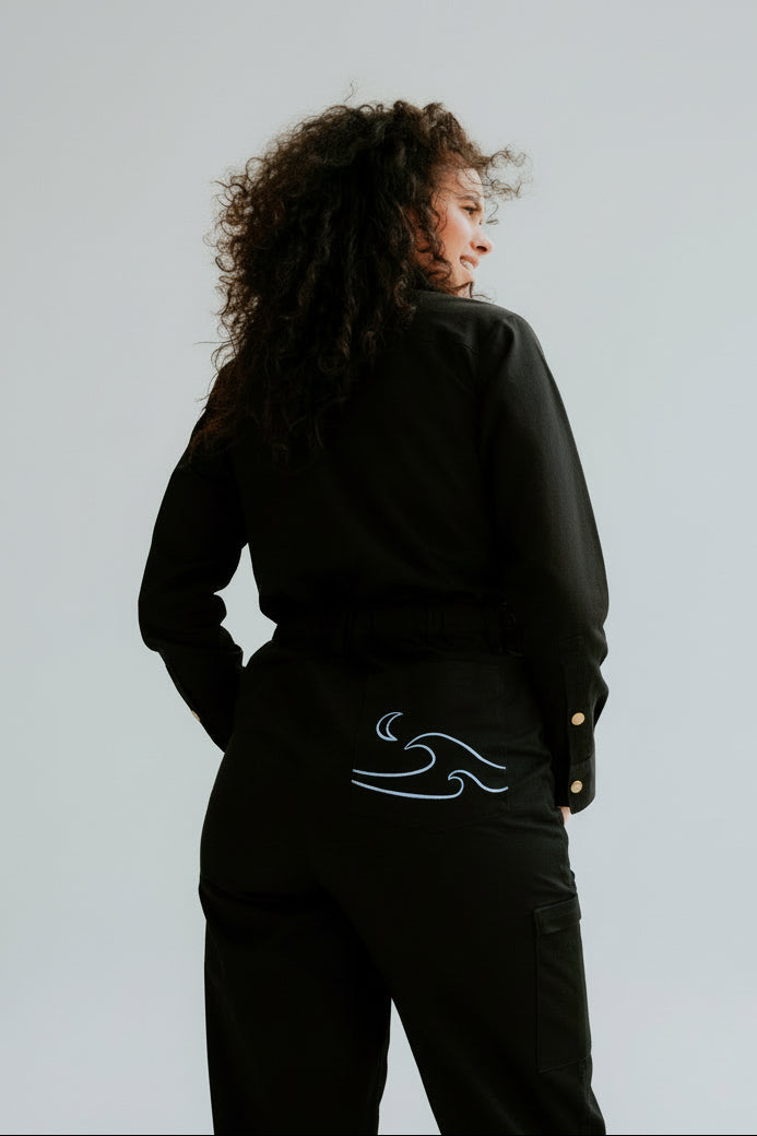 A model from behind wearing a pair of black women's coveralls with a blue embroidered wave design on the back pocket standing in front of a white background.