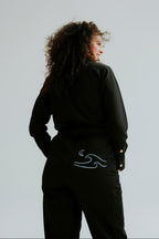 A model from behind wearing a pair of black women's coveralls with a blue embroidered wave design on the back pocket standing in front of a white background.