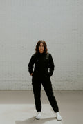 A model wearing a pair of black women's coveralls standing in front of a white brick wall. 