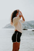 A woman standing by a body of water with mountains in the background wearing a pair of black women's board shorts with white piping.