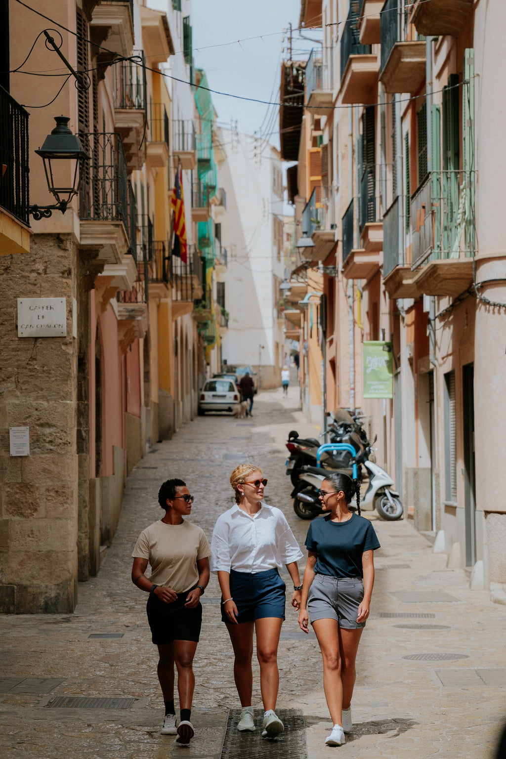 Three women walking down a narrow street in an urban area with buildings on either side wearing black, navy, and grey chino shorts. 