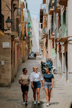 Three women walking down a narrow street in an urban area with buildings on either side wearing black, navy, and grey chino shorts. 