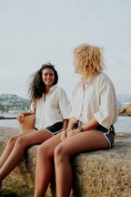 Two women sitting on a stone wall by the water, smiling and talking. Woman wearing board shorts / running shorts. Yachtwear. Women's deck shorts.