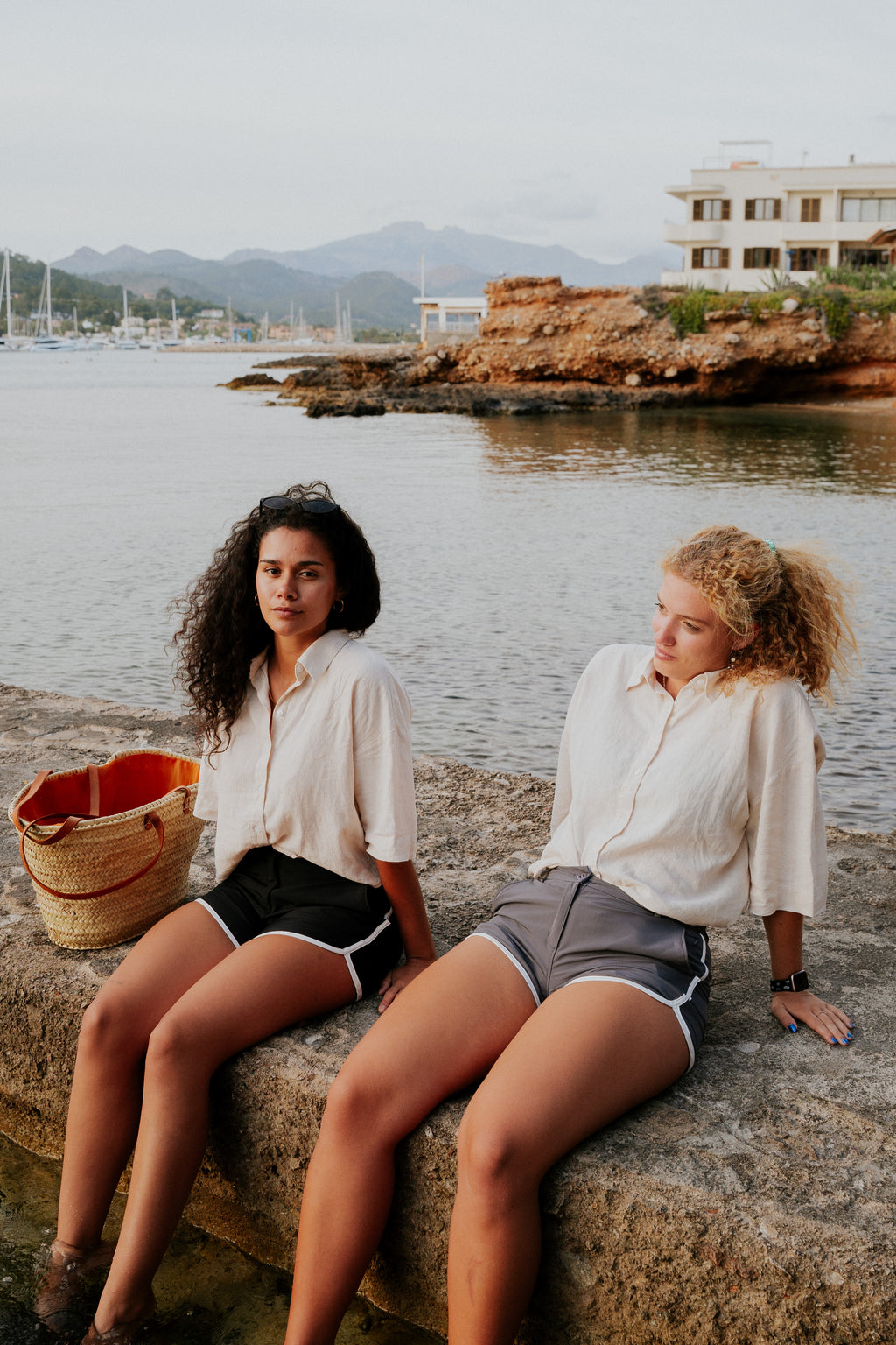 Two women sitting on a stone ledge by a waterfront with mountains in the background. Woman wearing board shorts / running shorts. Yachtwear. Women's deck shorts.