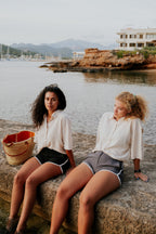 Two women sitting on a stone ledge by a waterfront with mountains in the background. Woman wearing board shorts / running shorts. Yachtwear. Women's deck shorts.