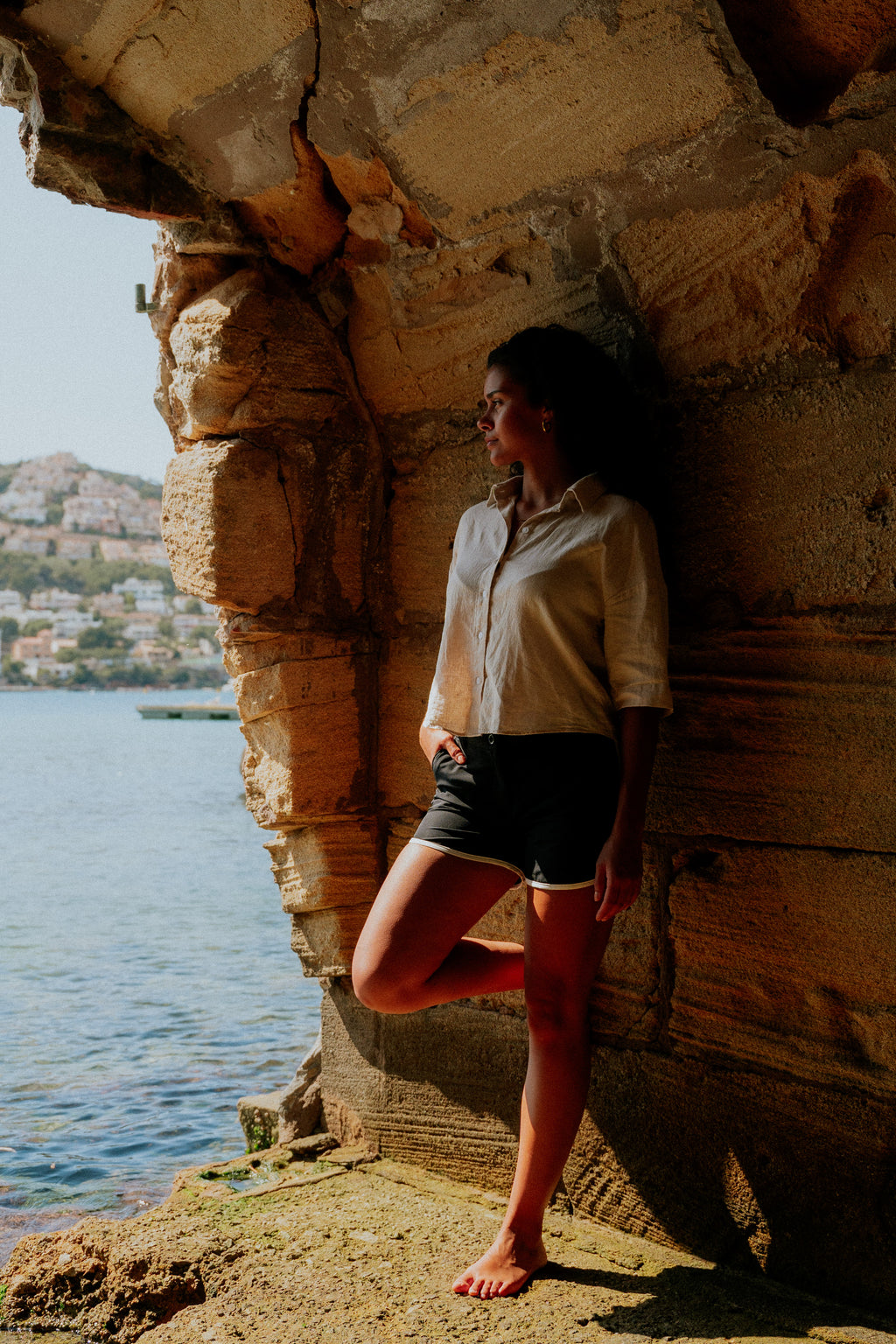 Woman standing against a stone wall by a body of water with a scenic background. Woman wearing board shorts / running shorts. Yachtwear. Women's deck shorts.
