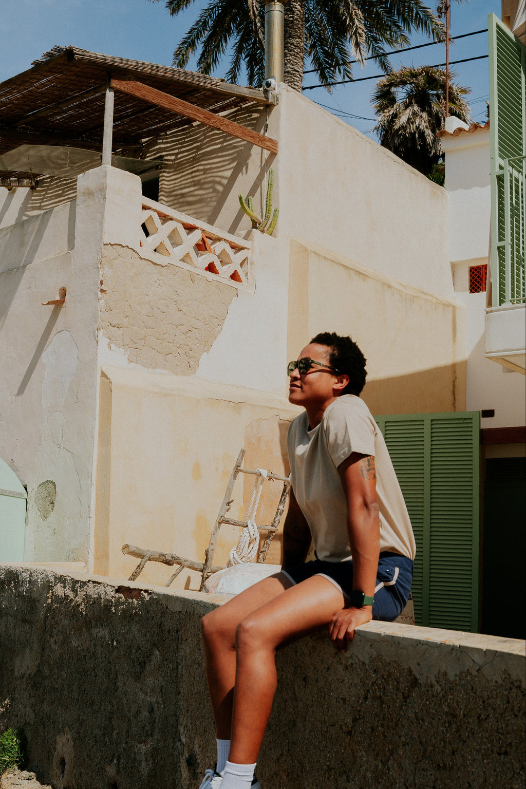 Person sitting on a ledge in a tropical setting with palm trees and a building in the background. Woman wearing board shorts / running shorts. Yachtwear. Women's deck shorts.