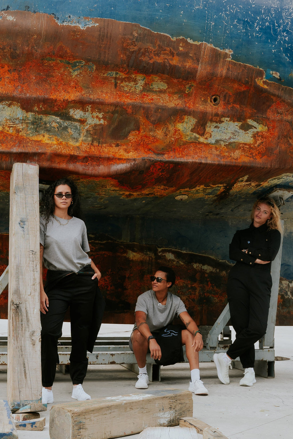 Three people posing in front of a large, rusted ship. Woman in black boiler suit / coveralls. Workwear, streetwear. Workwear style