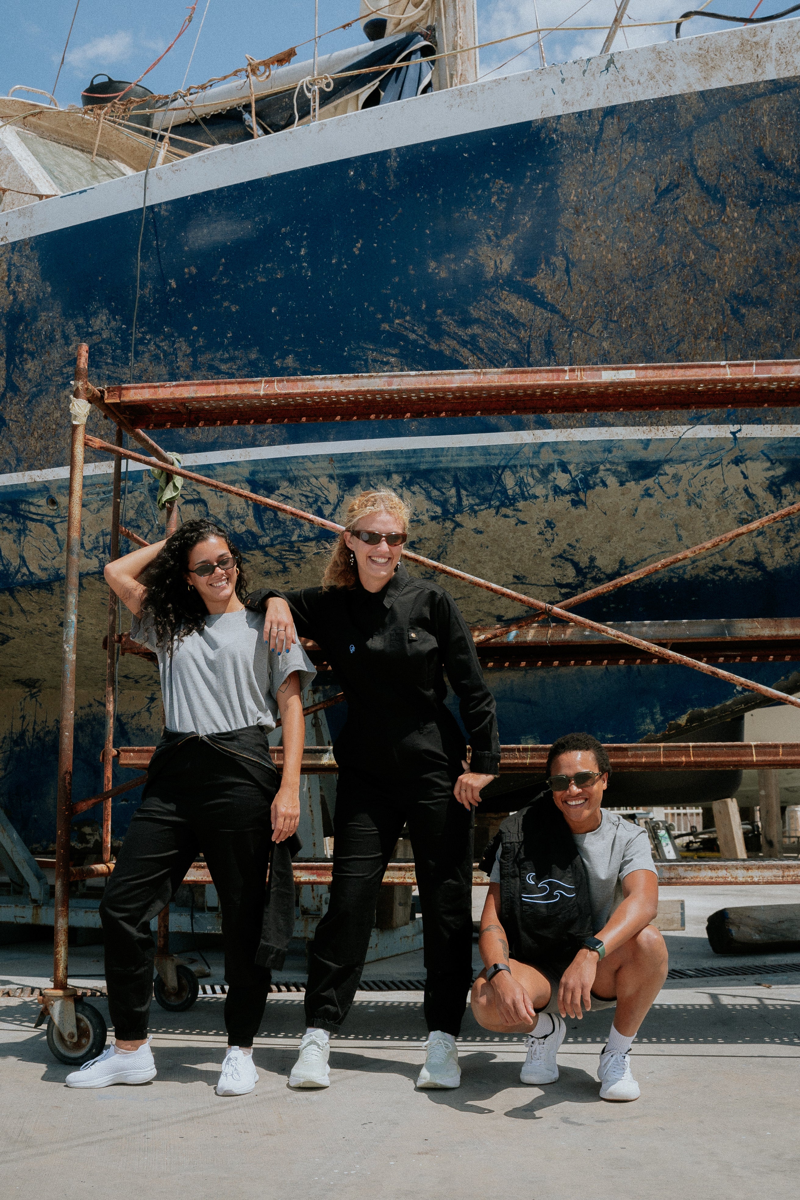 Three people posing in front of a large blue boat. Woman in black boiler suit / coveralls. Workwear, streetwear. Workwear style