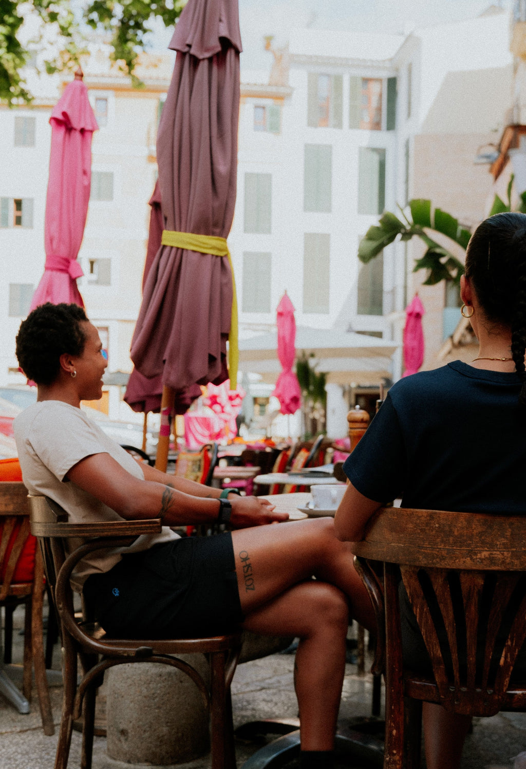 Two people sitting at an outdoor cafe table with umbrellas and buildings in the background. Woman wearing black chino shorts. Yachtwear. Women's Deck Shorts