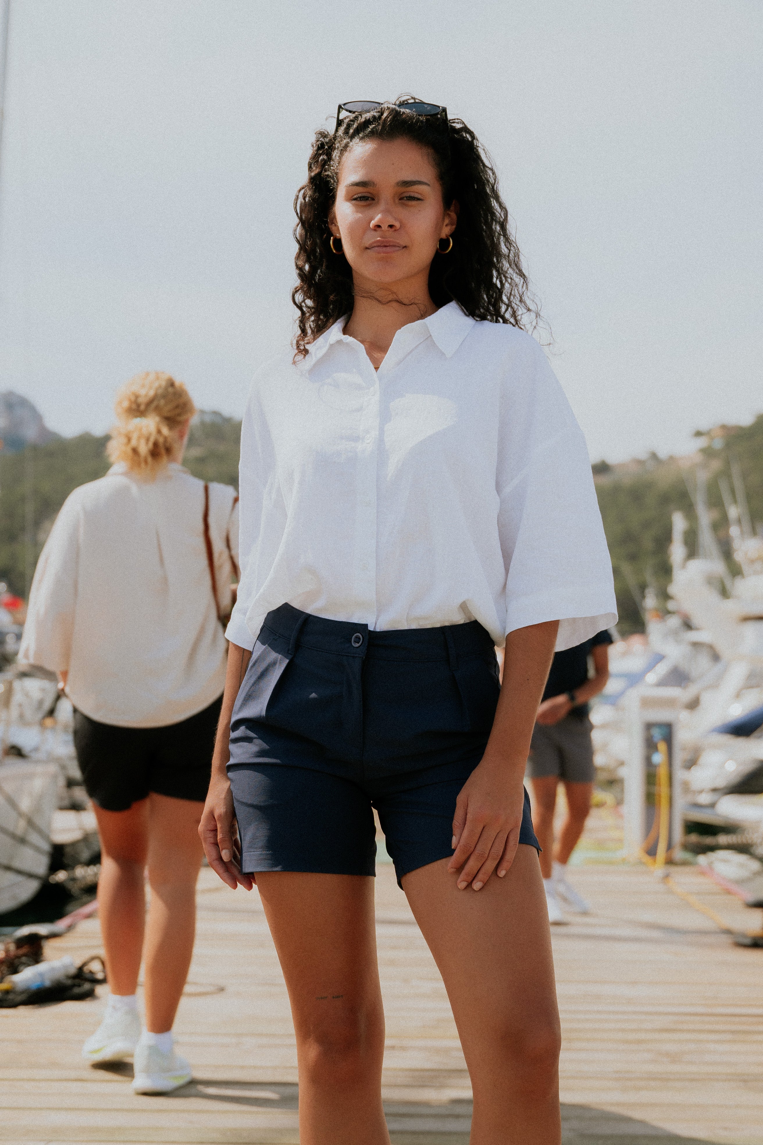 Woman in a white shirt and navy chino shorts standing on a dock with boats in the background. Deck Shorts. Yachtwear.