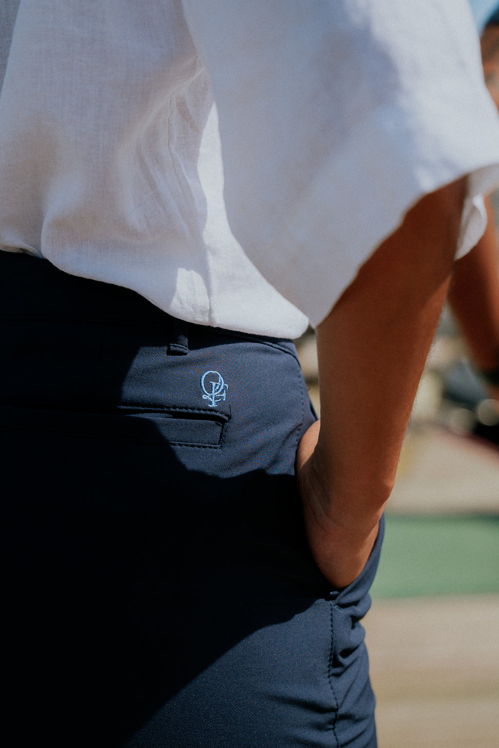 Person wearing a white shirt and navy chino shorts with Quay Figures' logo on the pocket, blurred background. Yachtwear. Women's deck shorts. 